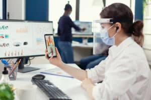 Businesswoman using wireless headphones during online conference on smartphone wearing face mask in workplace. Multiethnic coworkers working respecting social distance in business during global pandemic with coronavirus.