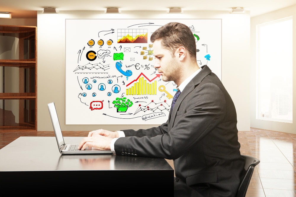 man typing in laptop with planning in white board beside