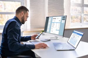 Business professional reviewing invoices on multiple screens while using a smartphone at a modern office desk