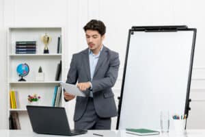 Businessman in suit presenting with clipboard beside blank whiteboard in modern office