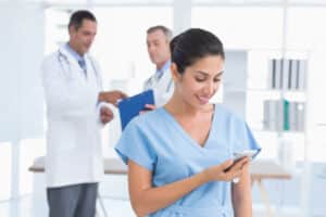 Smiling female nurse checking her smartphone while doctors discuss medical reports in the background at a modern clinic