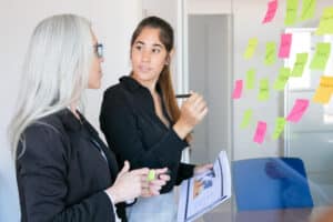 Two businesswomen discussing project strategy using sticky notes on glass wall