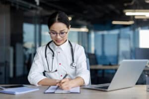 What Is a Scribe in Healthcare Smiling female doctor writing medical notes at desk with laptop