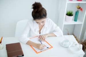 what is a medical scribe Young woman in white shirt writing on clipboard at desk