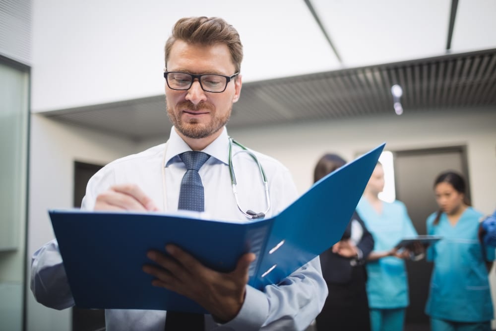 A focused male doctor reviews patient files while medical staff work in the background.