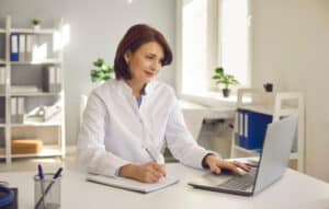 Female doctor in white coat working on laptop and taking notes
