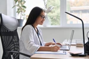 Female doctor in white coat working on laptop and taking notes at desk