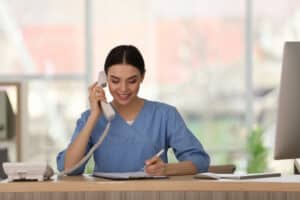 Medical receptionist in scrubs talking on phone and writing notes