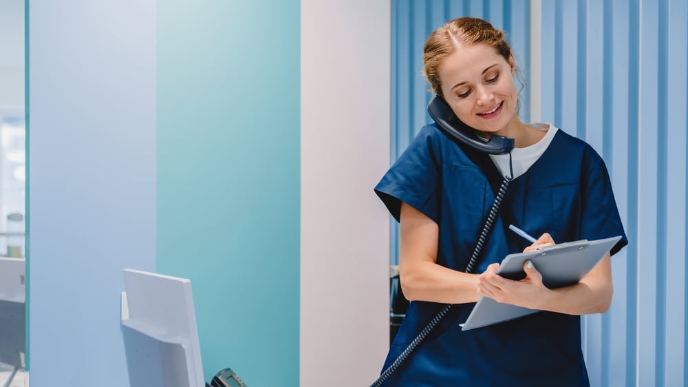 Female medical assistant holding a clipboard while writing and answering a phone call in a clinical setting.