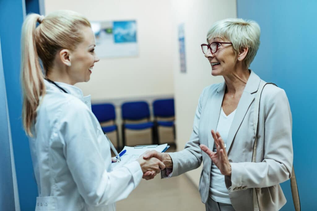 Happy senior woman shaking hands with a doctor while discussing care in a hospital hallway, specialty clinic referral follow-up workflow concept.