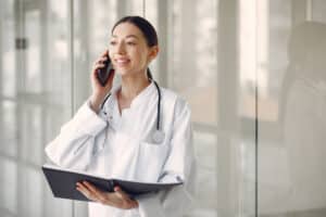Female doctor in a white medical uniform standing in a clinic hallway while talking on the phone, illustrating a patient callback system for clinics.