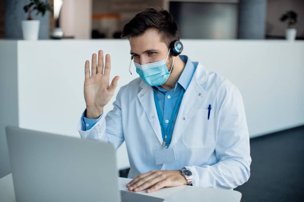 Male doctor waving during a video call on a laptop at a medical clinic, demonstrating a patient callback system for medical practices.