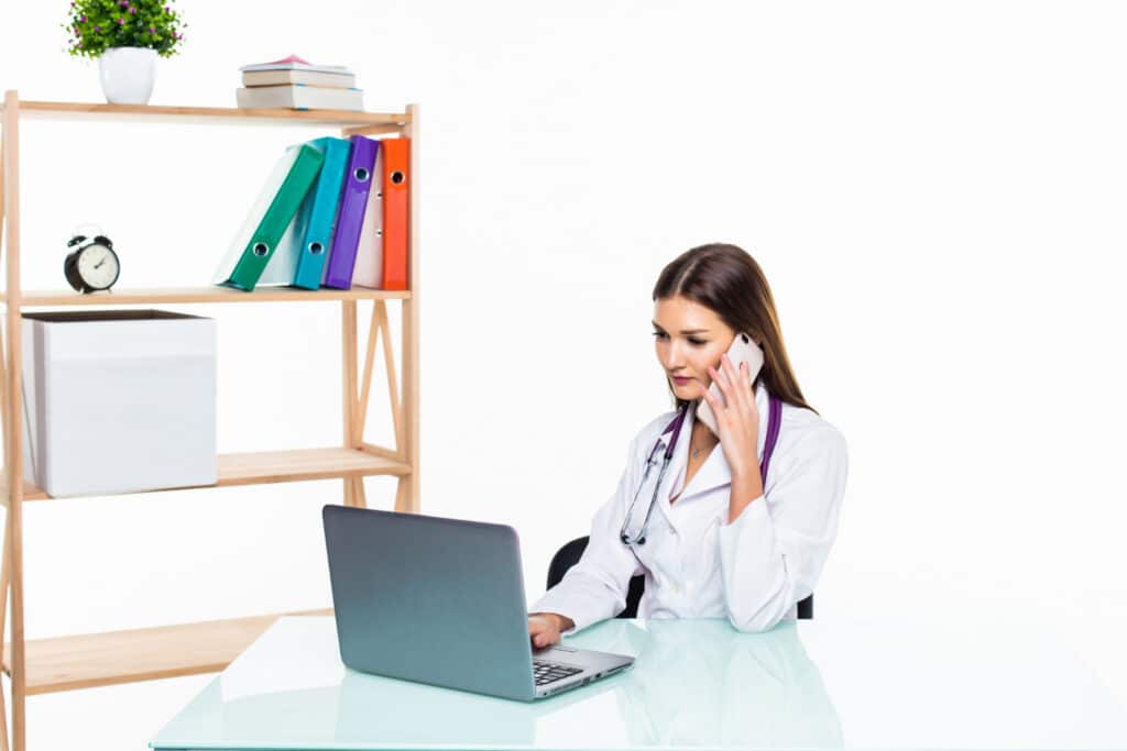 Virtual medical assistant support queues, female doctor sitting at a desk while speaking on the phone and working on a laptop during patient support tasks.