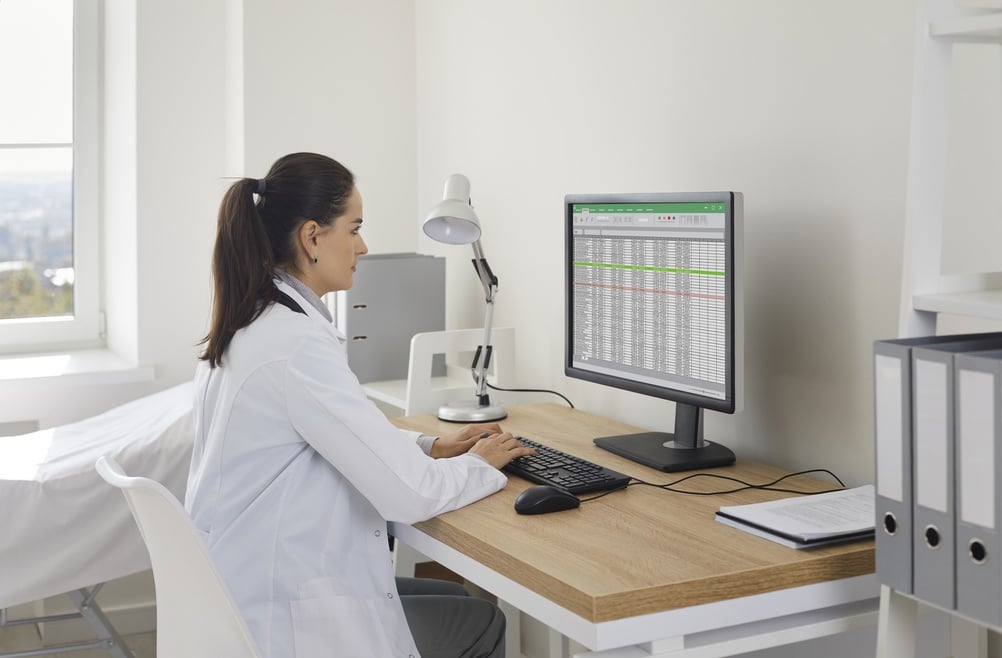 Female doctor in an office reviewing patient records and spreadsheets on a computer, highlighting medical operations and data management.