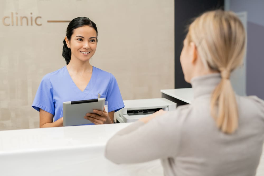 Happy young medical assistant holding a tablet and standing at a reception desk, representing efficient healthcare front desk customer service.
