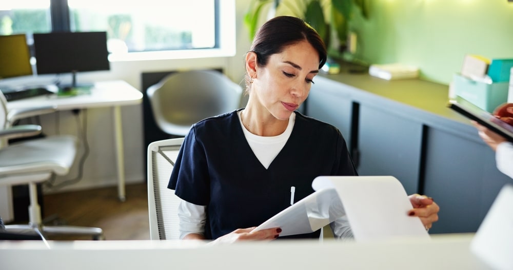 Healthcare receptionist helping a female patient complete medical forms and appointment paperwork at a clinic front desk