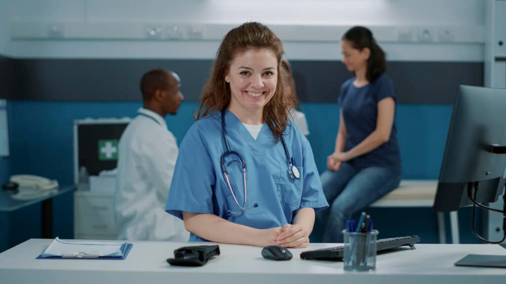 Smiling nurse in uniform sitting at an office desk, providing virtual medical assistant support for front desk operations and patient communication.