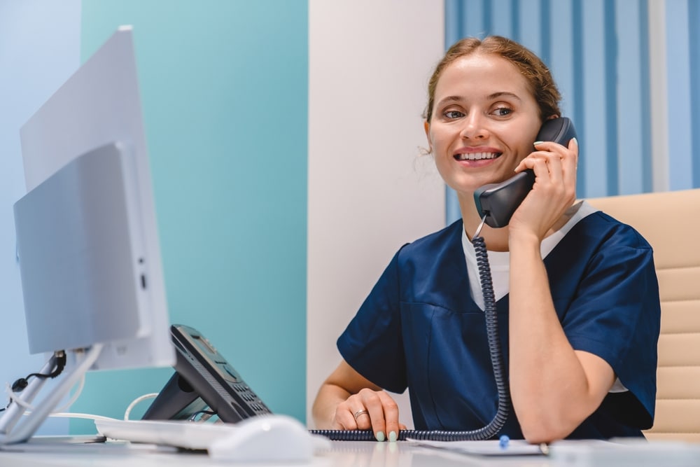 Smiling young female nurse receptionist talking on the phone at work, managing patient follow-up calls in a medical practice.