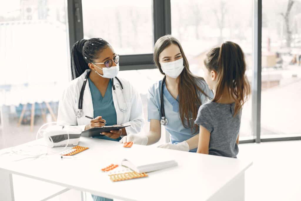 Two female doctors wearing masks talk to a young girl in a medical office.